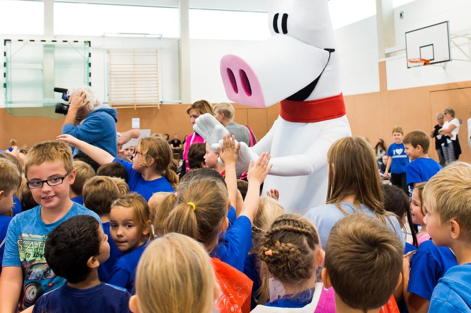 Ein großer, weißer Stofftier-Charakter interagiert mit aufgeregten Kindern in blauen T-Shirts in einer Turnhalle.
